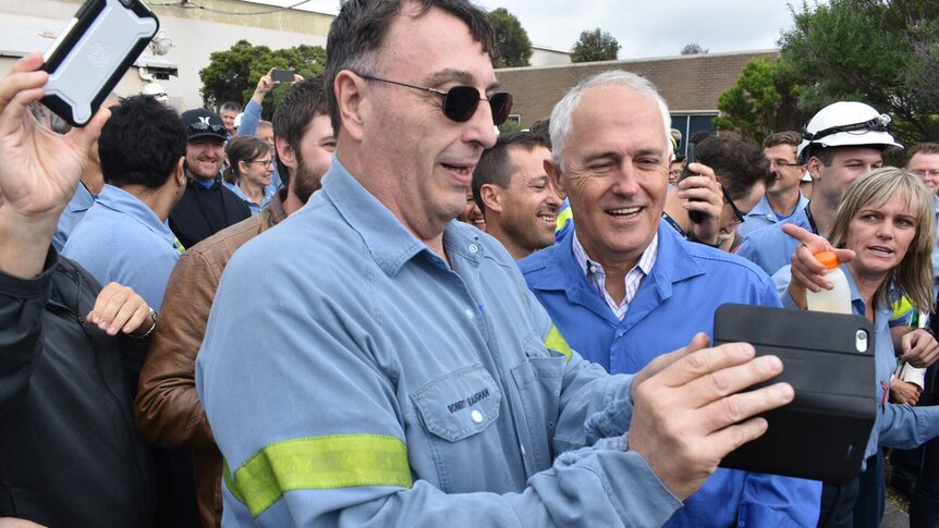 A man in a blue shirt and glasses holds a mobile phone next to Prime Minister Malcolm Turnbull, with a crowd behind.