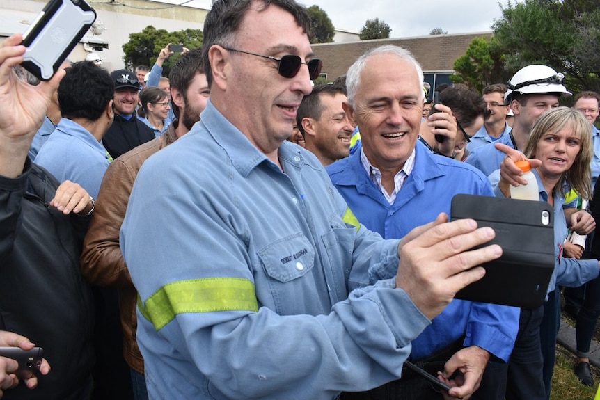 A man in a blue shirt and glasses holds a mobile phone next to Prime Minister Malcolm Turnbull, with a crowd behind.