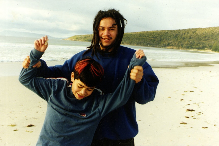 Two boys stands on a coast. The elder is lifting his brother's hands high and smiling for the camera. 