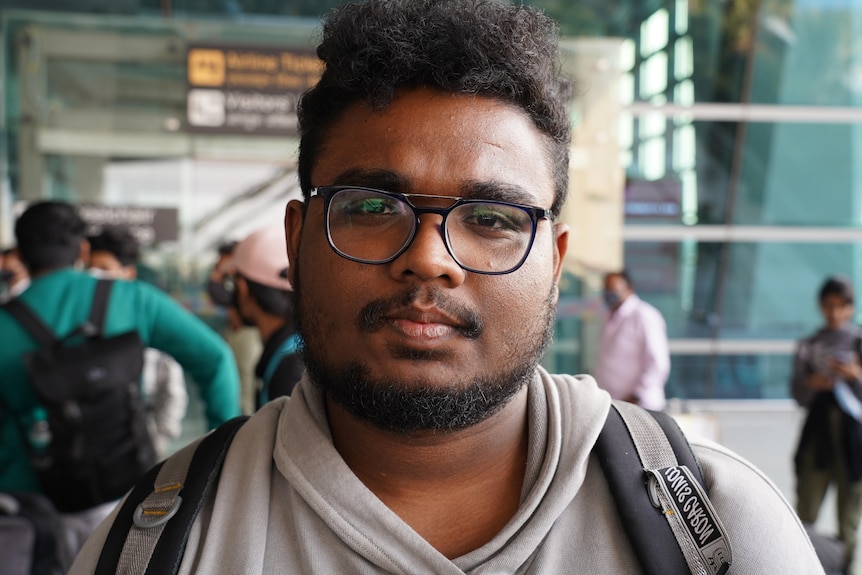 A young Indian man in a cream hoodie, black glasses, and a backpack stands at an airport