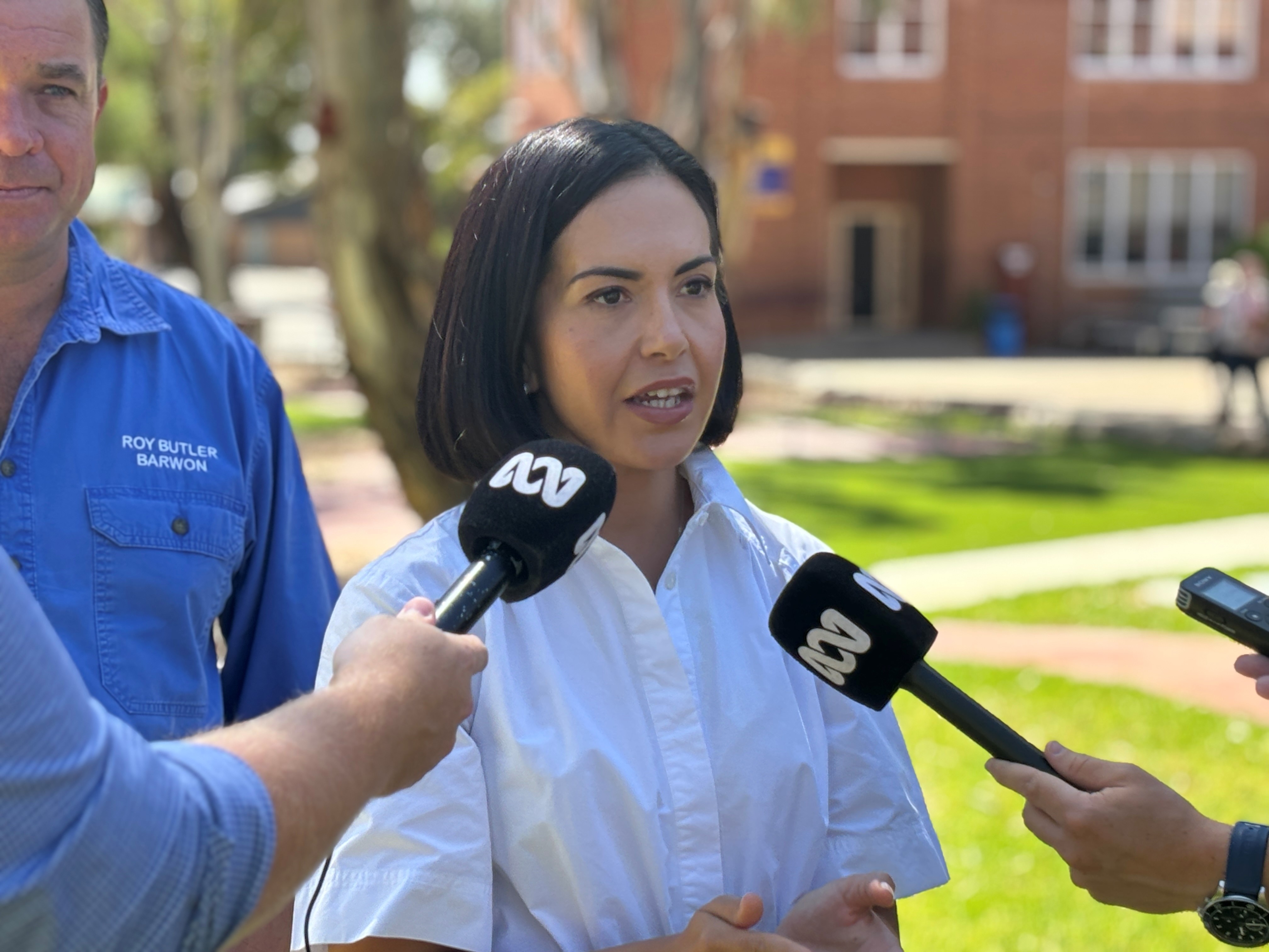 A woman with black hair in a white dress speaking to a group of microphones in front of a grass patch and group of buildings
