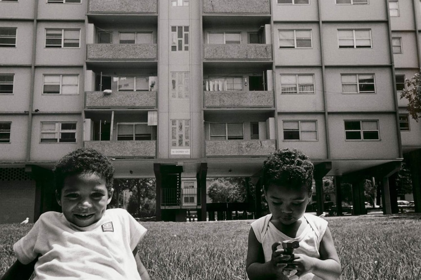 Shows two young brothers, seated on the grass, outside the low-rise public housing