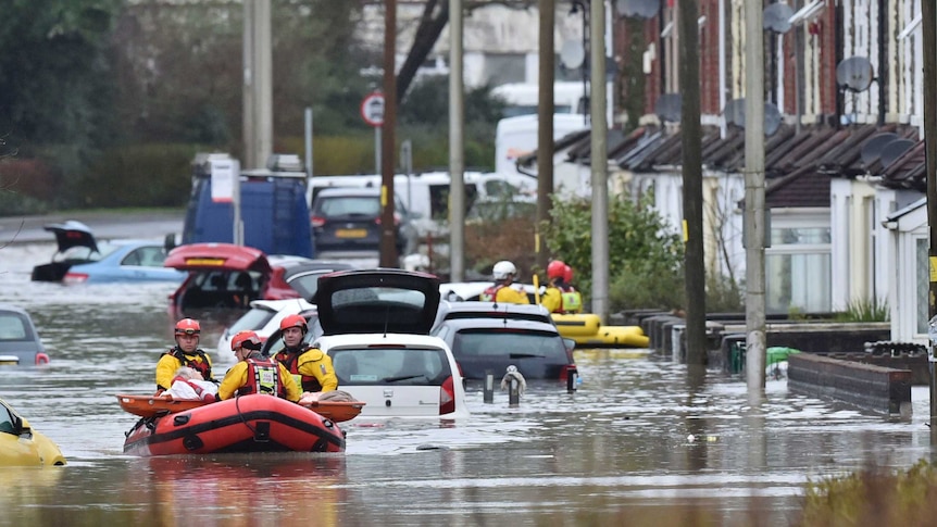 In a flooded street in a town, rescuers dressed in yellow ferry a local resident in a inflatable raft next to houses