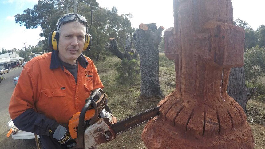 A man in a hi-viz shirt wearing earmuffs wields a chainsaw which he is using to carve a tree into a cross-shaped chess piece.
