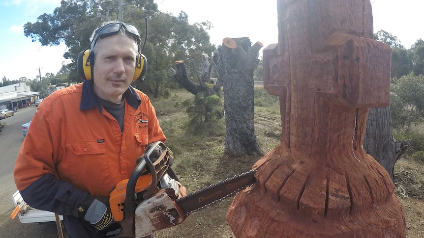 A man in a hi-viz shirt wearing earmuffs wields a chainsaw which he is using to carve a tree into a cross-shaped chess piece.
