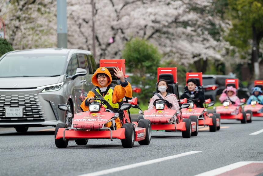 An Asian man in a costume waves to camera at the head of a line of go karts on cherry blossom-lined street