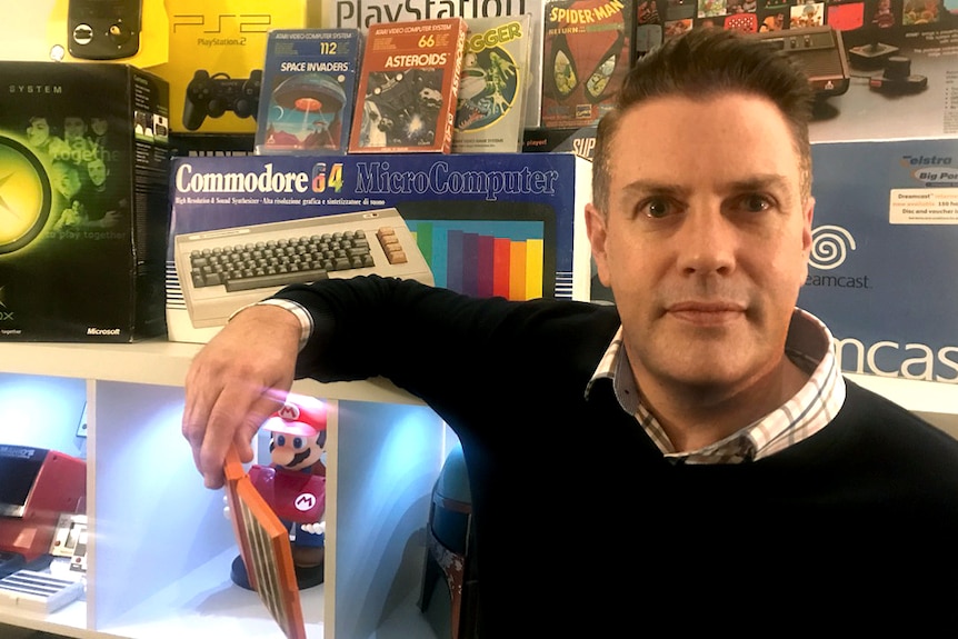 A man in a black top stands with a collection of retro computer games and equipment.