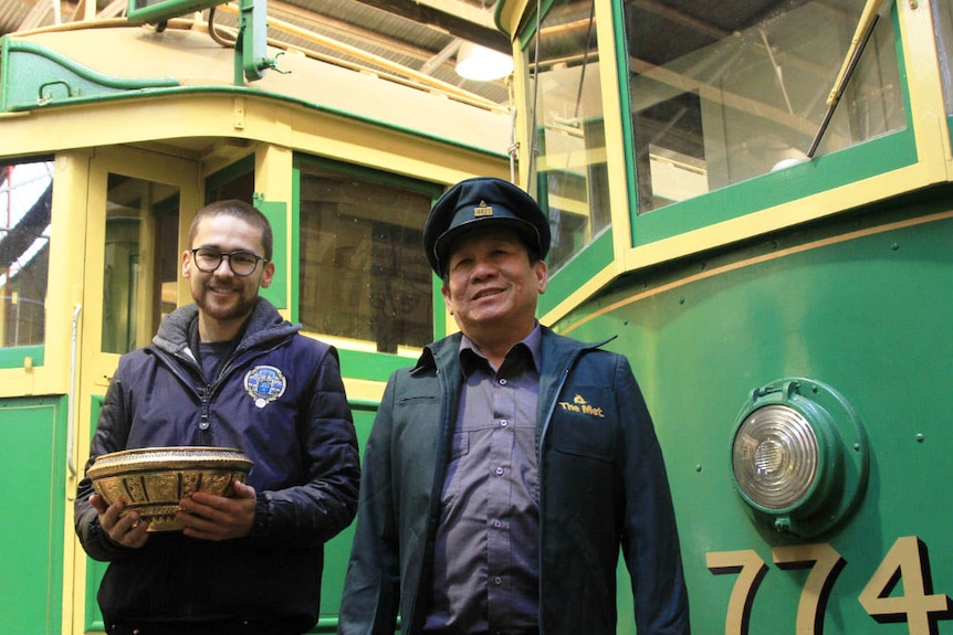 Two men smiling standing in front of two yellow and green trams. One holds a golden metal bowl.