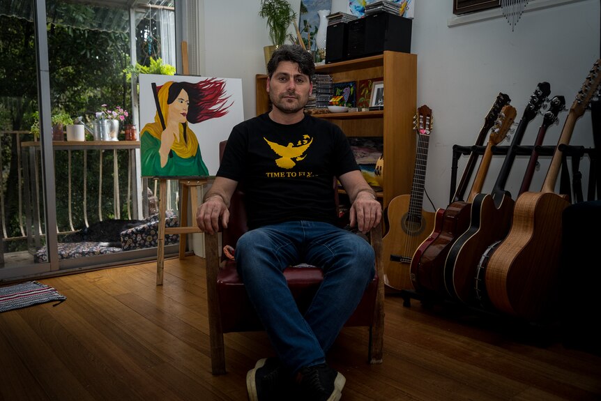 A man wearing a t-shirt sits on a chair, surrounded by guitars and a painting
