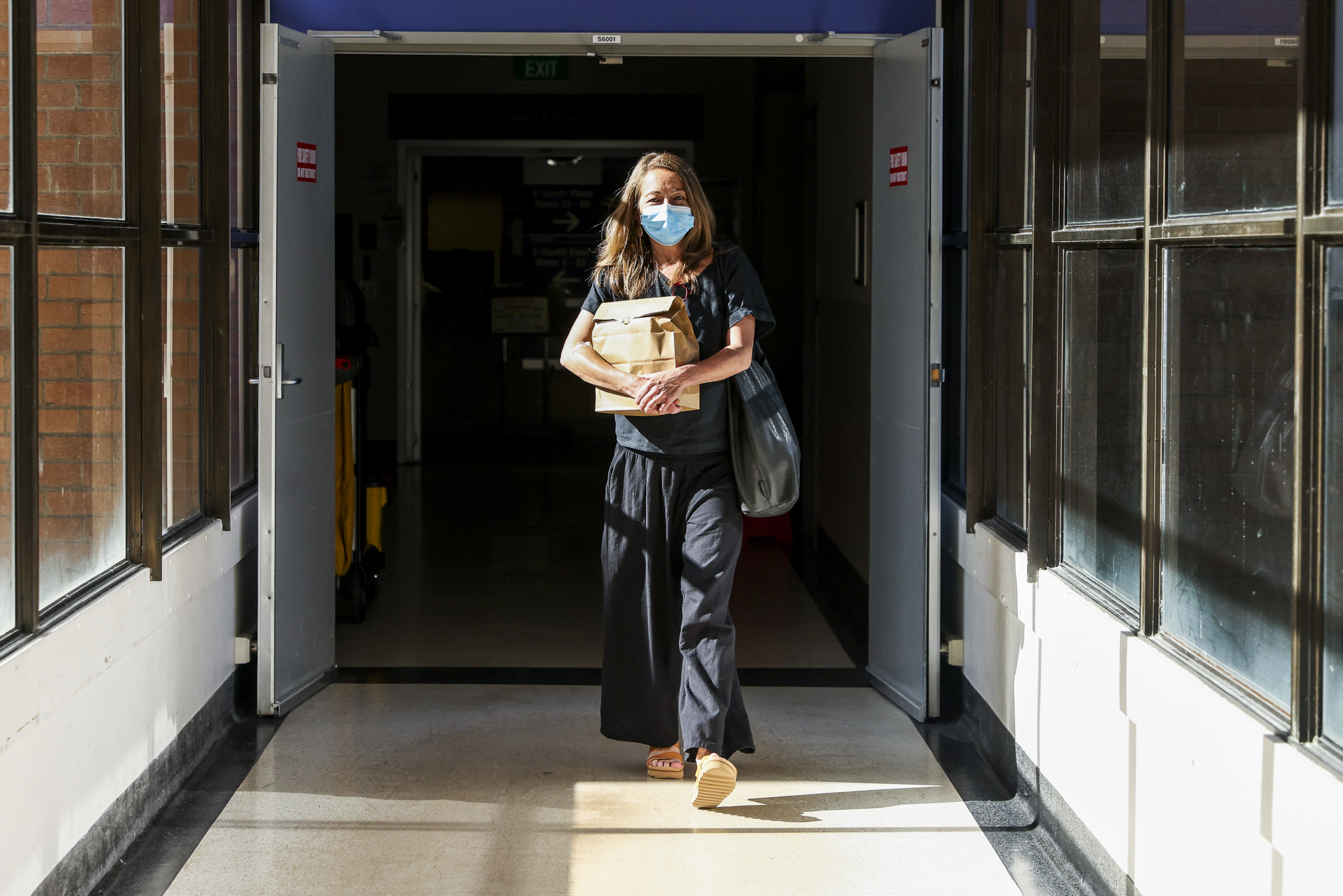 A woman with a mask on walks down a hospital corridor with a large paper bag of medication.