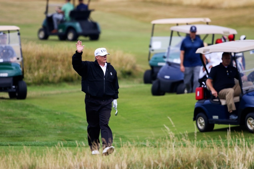 Donald Trump waves at protesters as he walks on a golf course surrounded by golf carts