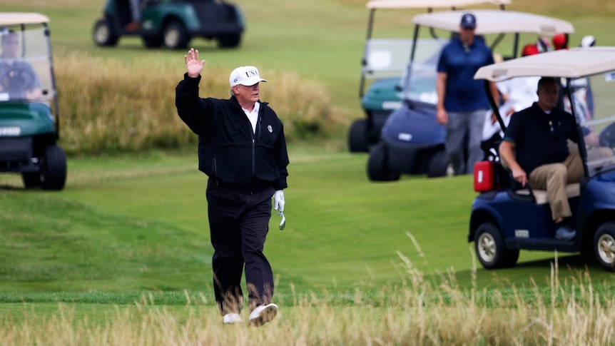 Donald Trump waves at protesters as he walks on a golf course surrounded by golf carts