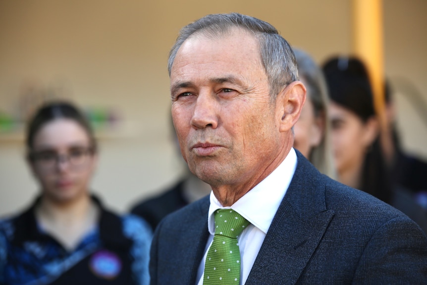 A medium close-up shot of WA Premier Roger Cook wearing a dark suit, white shirt and green tie, with people in the background.