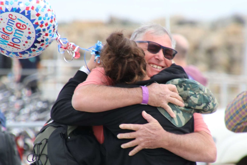 A man and daughter embrace and smile on a wharf while she holds a colourful balloon that says "welcome home".