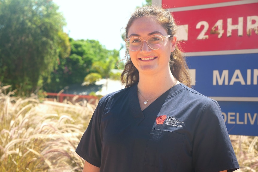 A lady with glasses in medical scrubs standing in front of a hospital sign outside. 