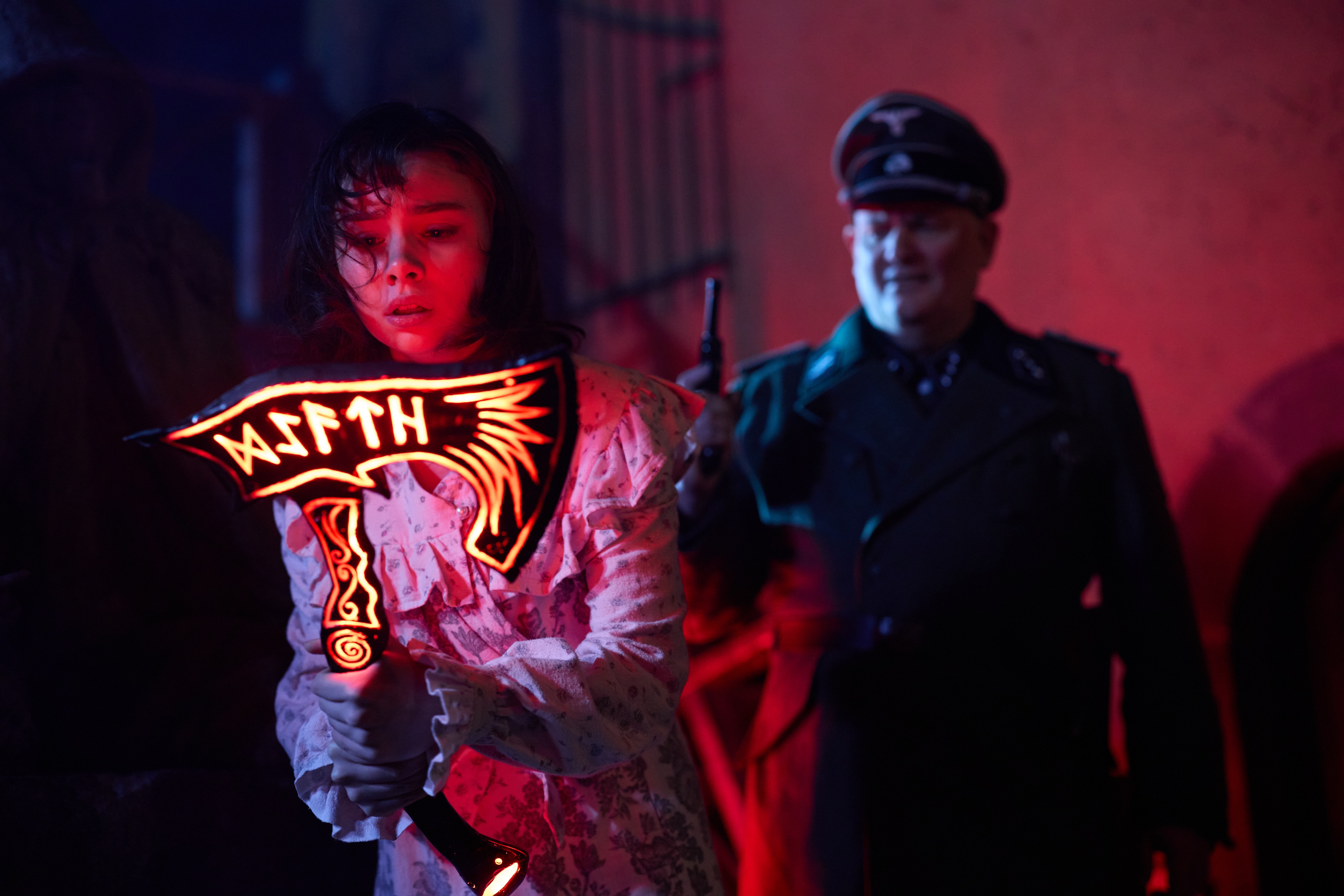 A nazi points a gun at a young girl holding a sythe
