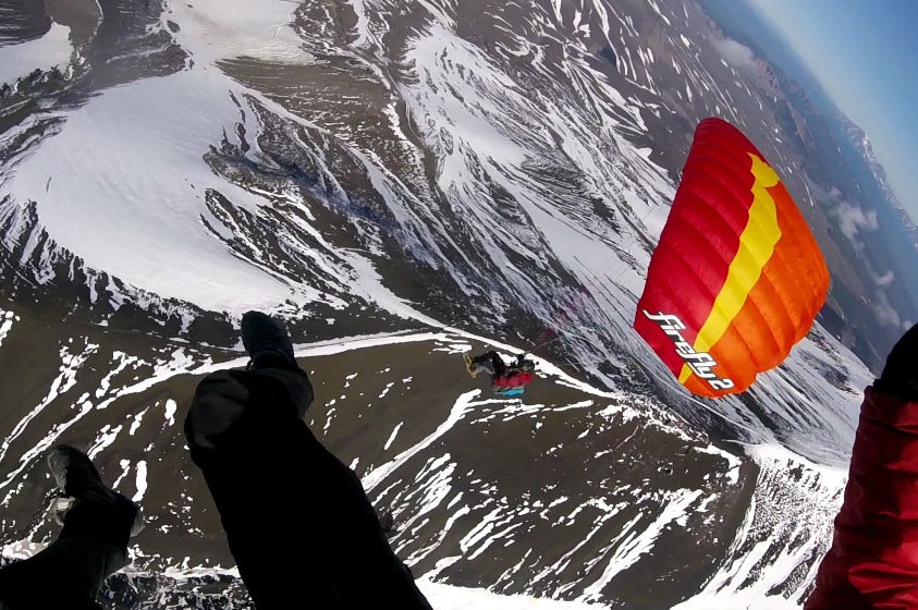 A speed flyer looks down over another pilot's parachute soaring above snow-covered mountains.