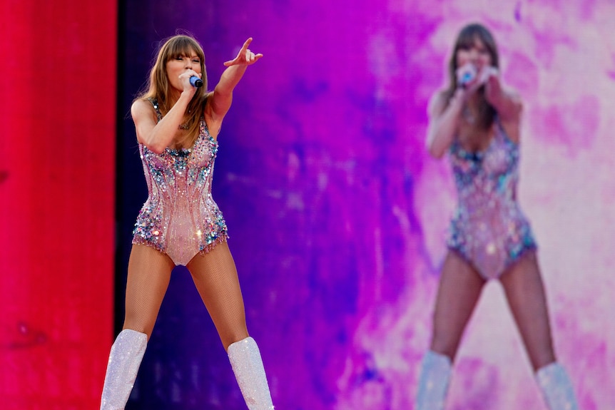 Taylor Swift dressed in a sparkly leotard points her finger on stage as she sings into a microphone