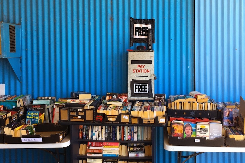 A pay station that has a 'Free' sign sits on top of a table of books, in front of a blue steel shed.