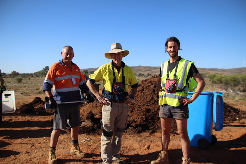 Three men smiling in high vis clothing standing in front of a large pile of compost