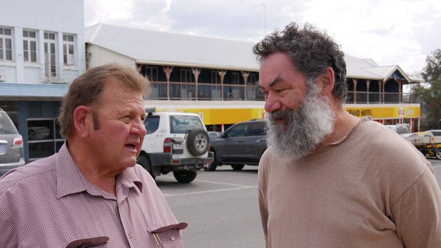 Two middle aged men talk to each other in a street. Man on left has a red checked shirt and the other man wears a plain t-shirt