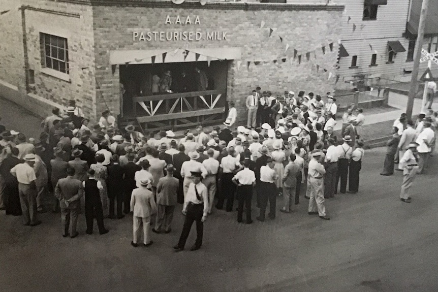 about 50 people gather outside a brick milk factory building