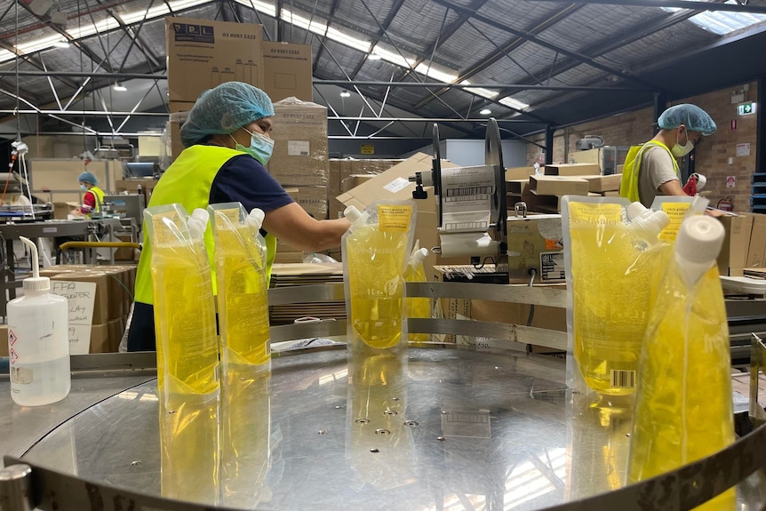 Cinco bolsas llenas de líquido amarillo para lavarse las manos se encuentran en una mesa giratoria con un trabajador empacandolas en una caja. 