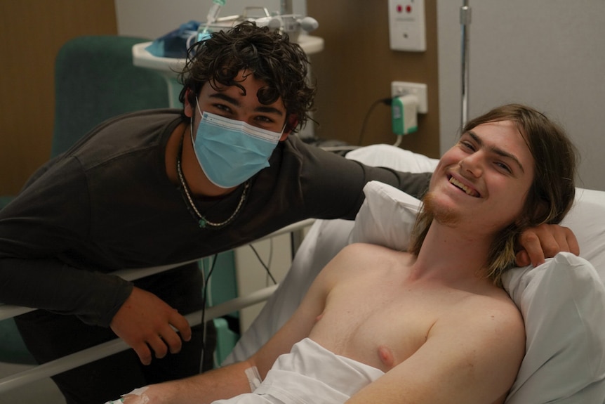 Two male teenagers, one in a hospital bed.