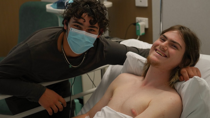 Two male teenagers, one in a hospital bed.