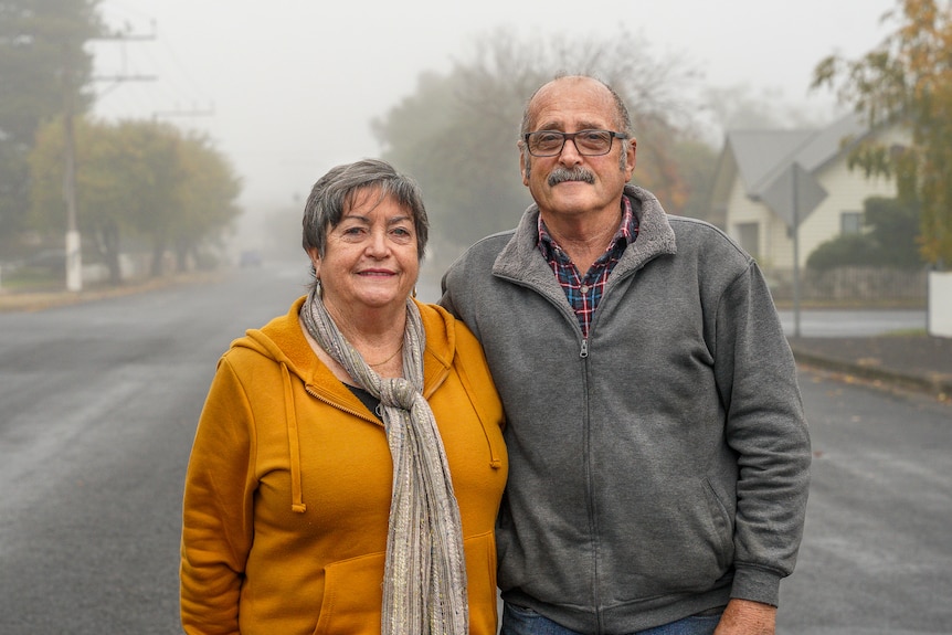 An older man and woman stand for a photo in a foggy street with their arms around each other.