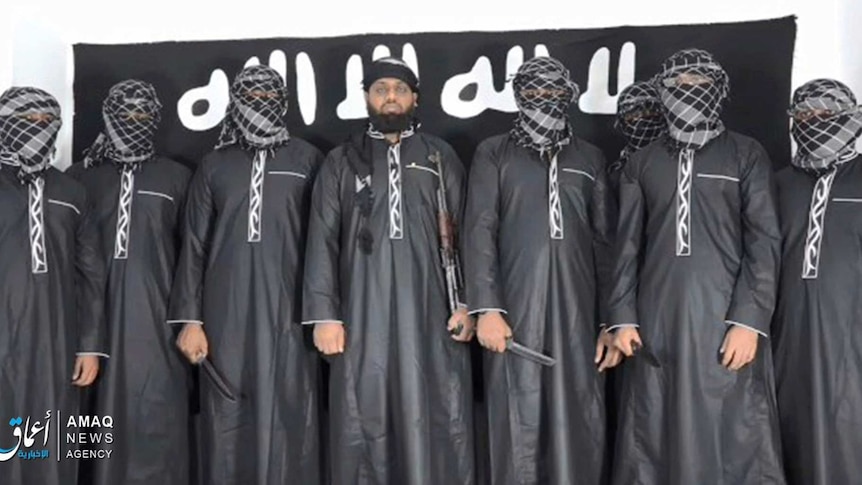 Eight men stand in a line in black robes, some holding knives, one, centre, is unmasked, standing in front of black IS flag.