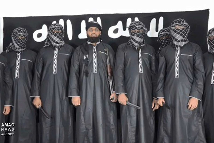Eight men stand in a line in black robes, some holding knives, one, centre, is unmasked, standing in front of black IS flag.