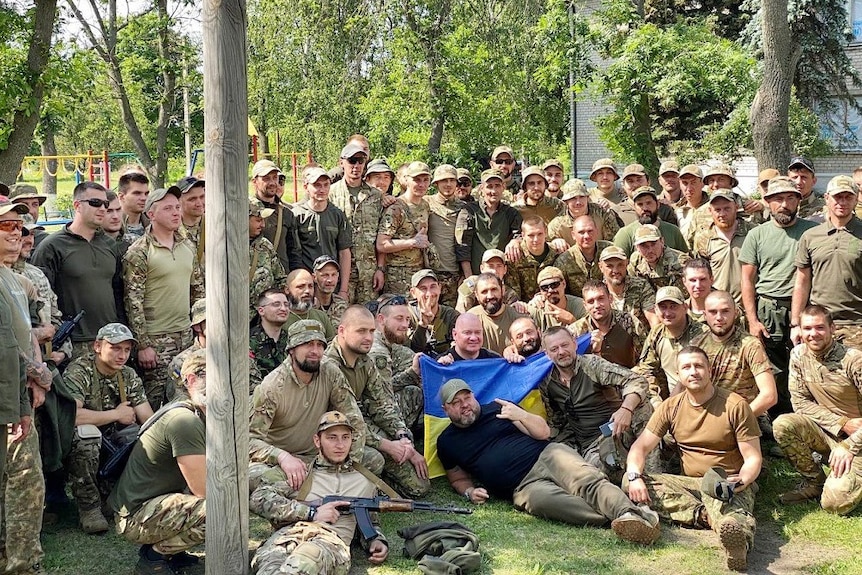 Two comedians and a large group of soldiers pose for a photo outdoors with the Ukrainian flag