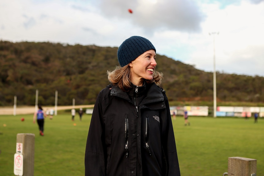 Woman wearing black jacket and beanie smiles with football ground in background