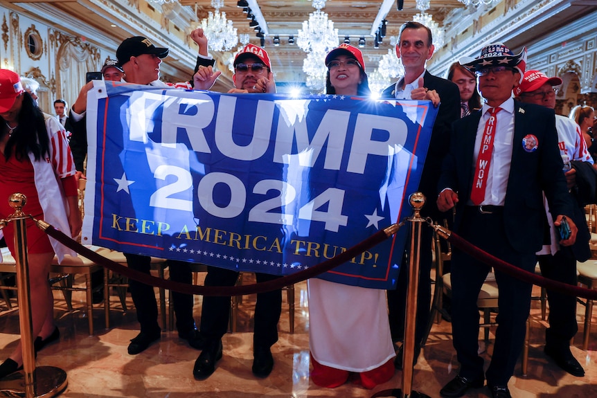 Supports of Donald Trump hold up a banner saying Trump 2024 inside a ballroom.