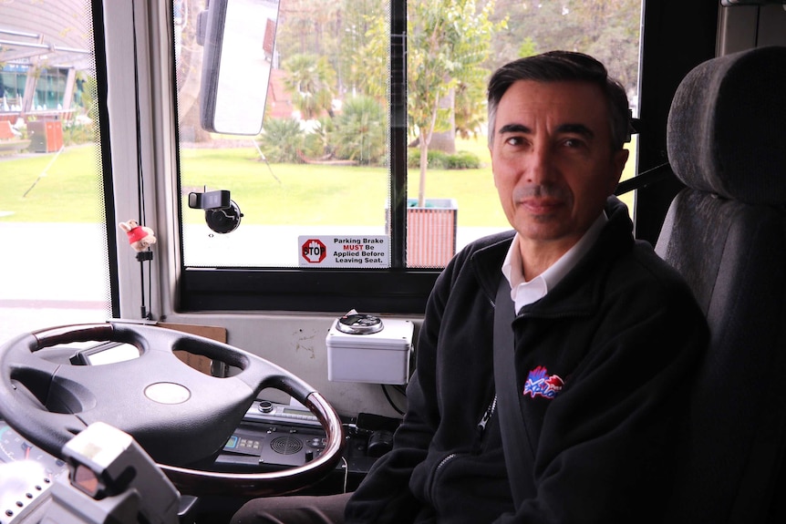 A bus driver wearing a seatbelt sits at the wheel of his stationary vehicle.
