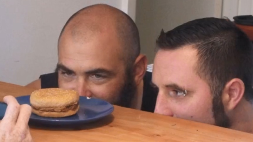 Two men stare at a burger sitting on a bench.