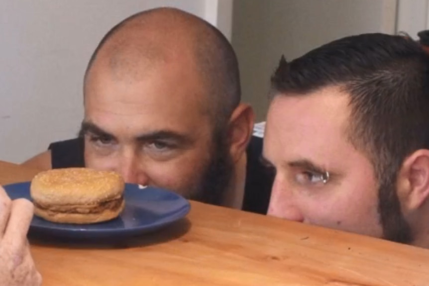 Two men stare at a burger sitting on a bench.