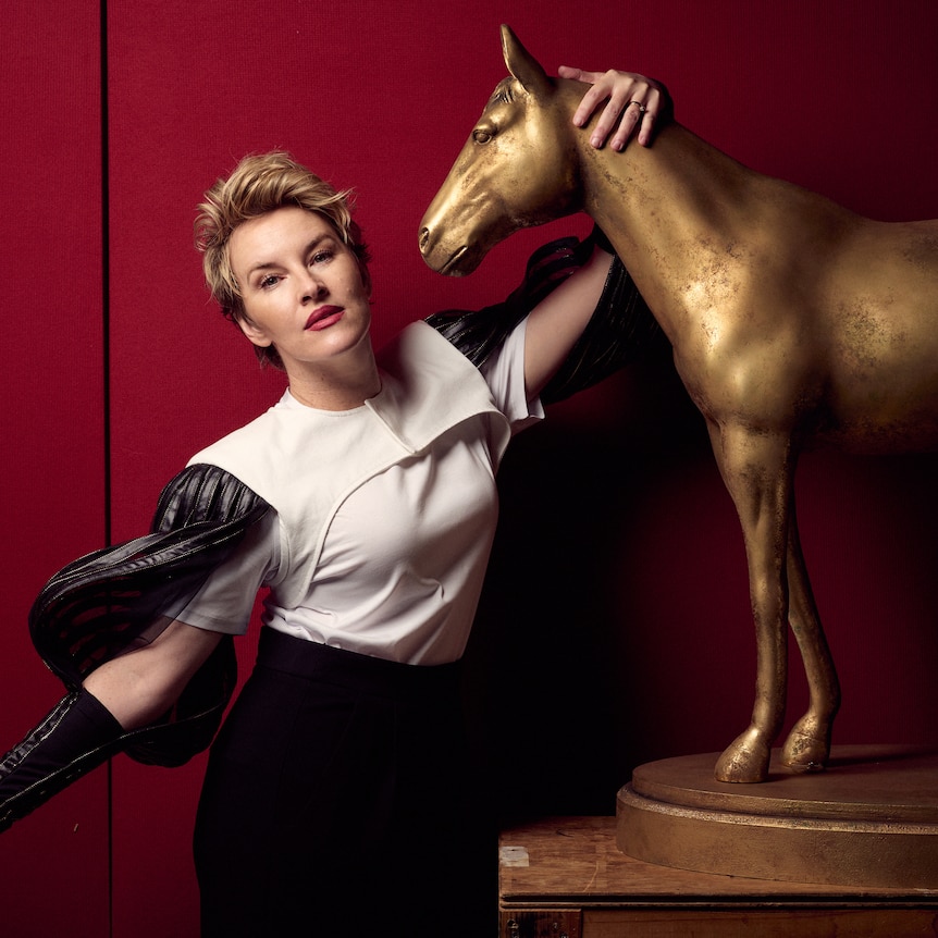 A blonde, short-haired woman in her 40s, looks into the camera lens. She has one arm around a bronze horse sculpture.