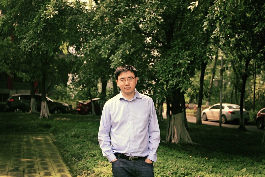 A man in a blue checked shirt stands in front of green trees.