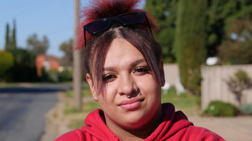 A young woman stands on a suburban street. She's wearing a red jumper and sunglasses on her head. 