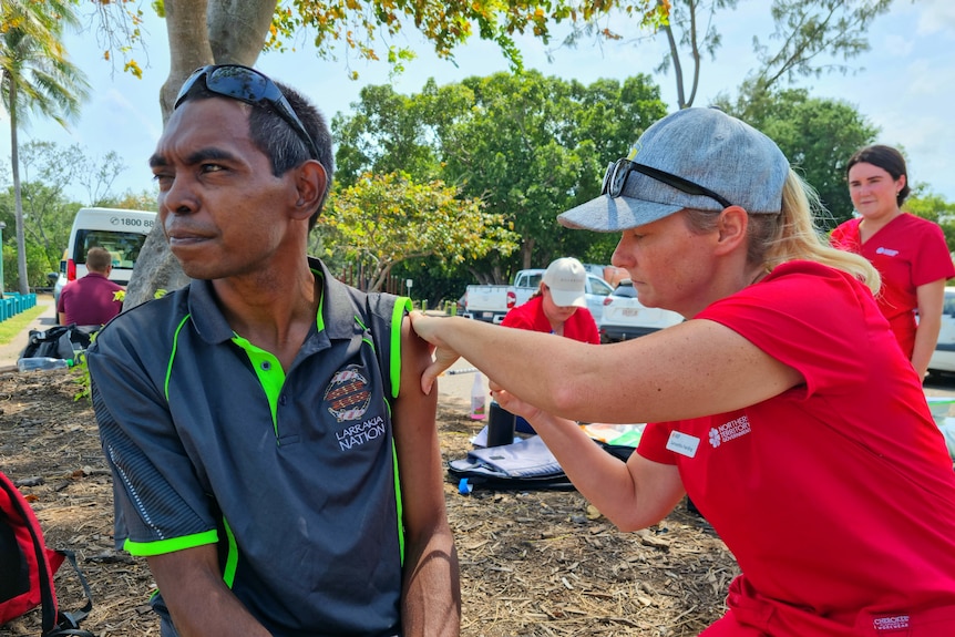 A woman injects a man with a vaccine at a park, with  greenery in the background. 