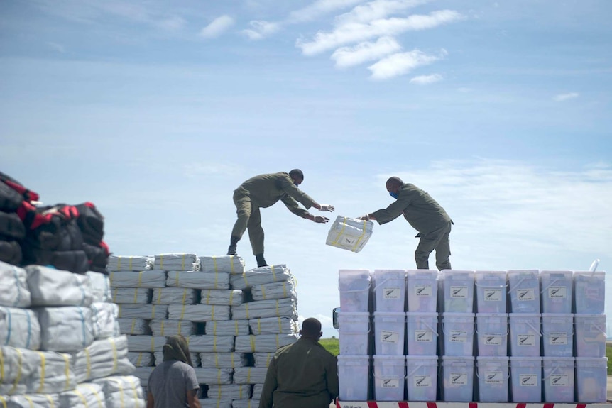 One man stands on a supply crate throwing supplies to another man on another crate.