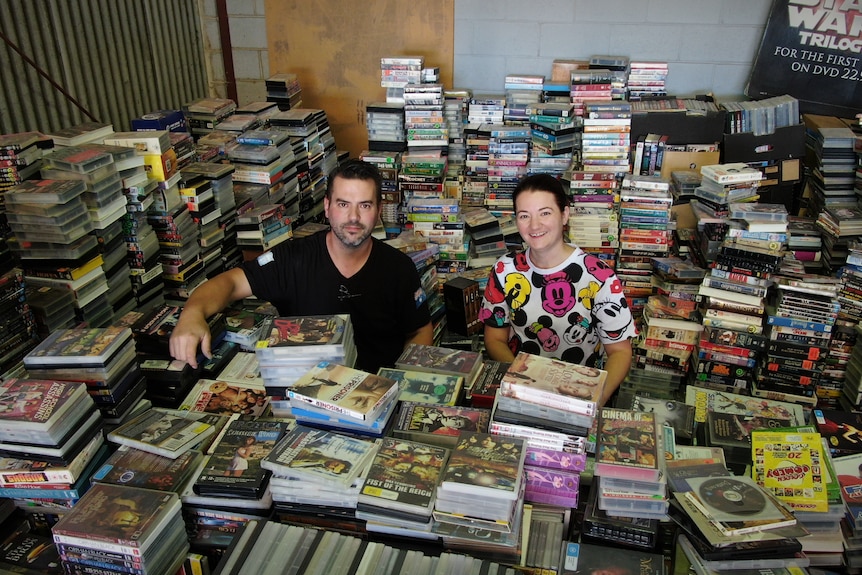 A man in a black shirt and a woman in a colourful mickey mouse t shirt stand amongst piles of video tapes