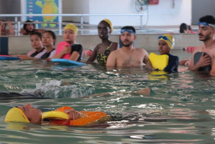 A woman in orange and yellow cap floats on her back in a swimming pool as a group of students watch on.