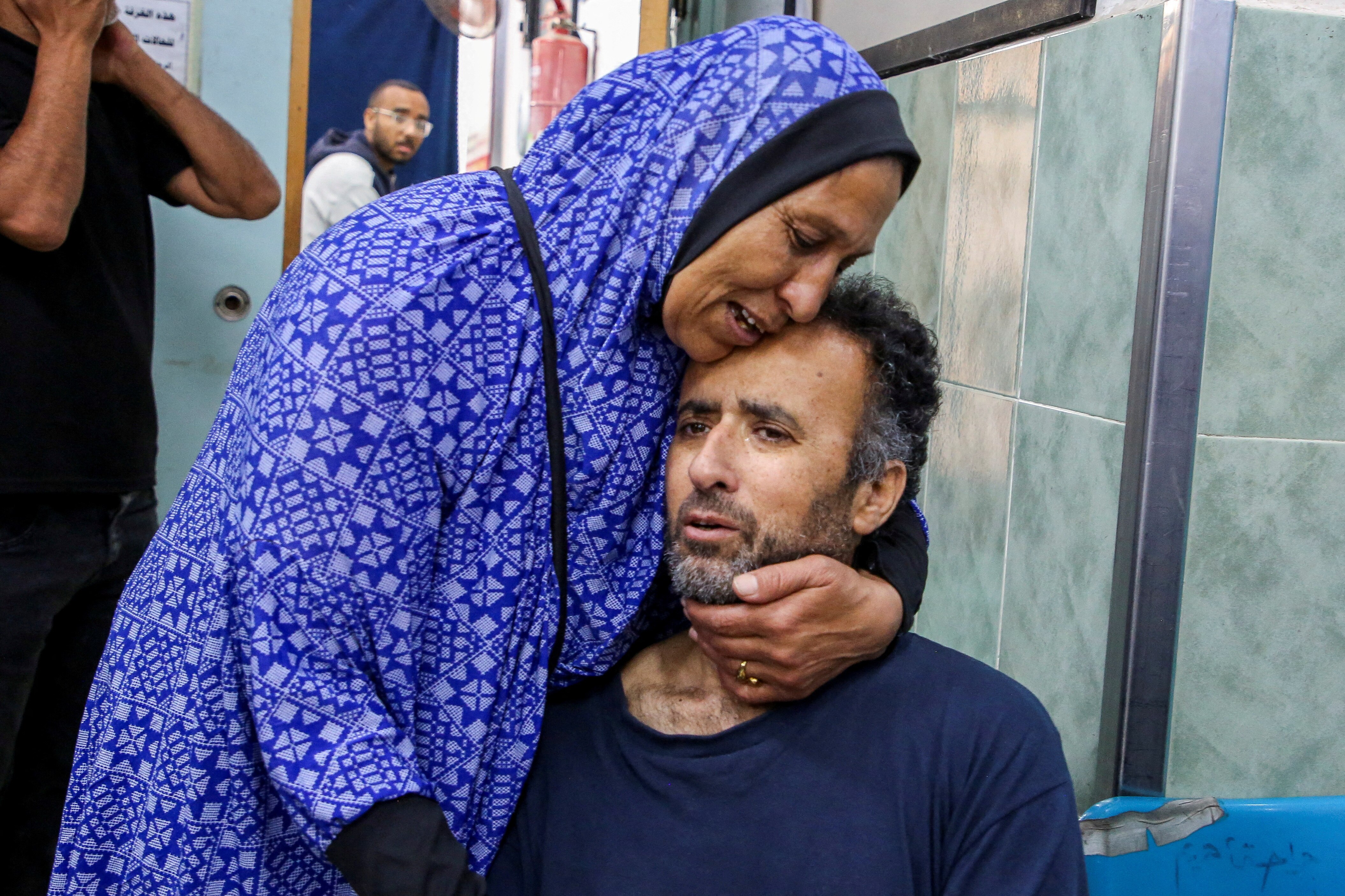 A middle-aged Palestinian man seated in a blue T-shirt stares straight ahead as he is hugged by a crying woman.