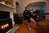 A woman dressed in pink uggs and a red beanie sits on a dining chair next to a heater in the living room.