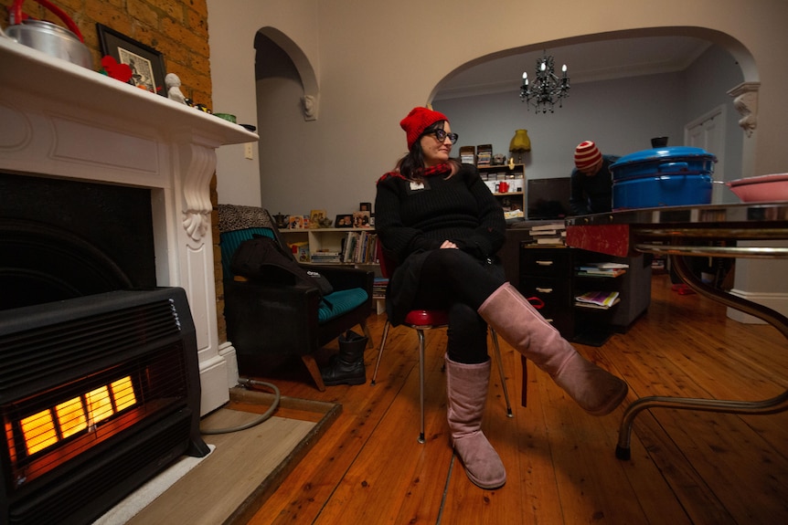 A woman dressed in pink uggs and a red beanie sits on a dining chair next to a heater in the living room.
