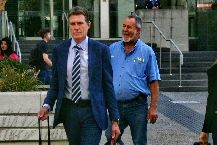 Two men, one in a suit and the other in a blue collar work shirt, walk along a street. 