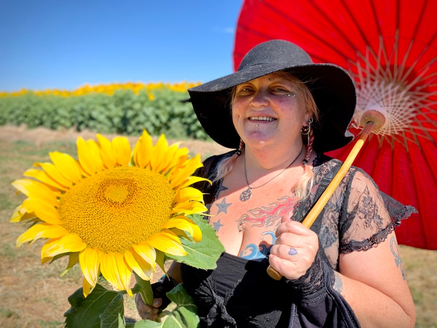 A woman with a black hat and umbrella is holing a large sunflower